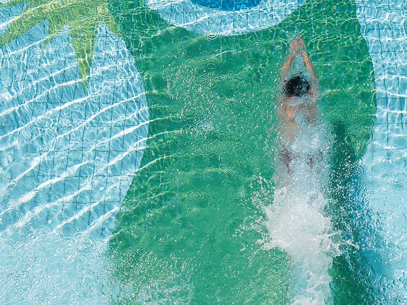 Ein Schwimmer taucht durch das klare Wasser eines Pools, während sich Wellen um ihn herum ausbreiten. Von oben betrachtet, ist ein farbenfrohes Muster auf dem Beckenboden zu sehen, welches das Logo des Sonnenguts zeigt