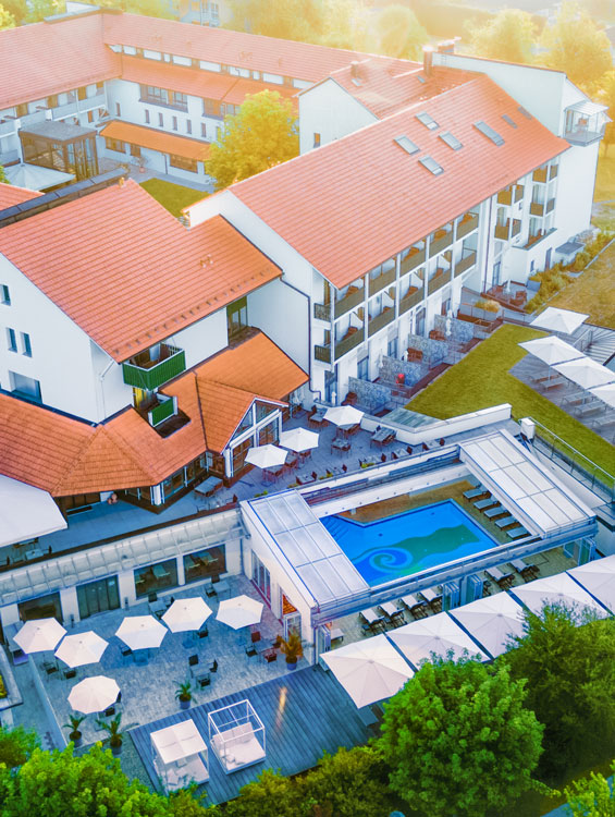 Eine Luftaufnahme des Hotel Sonnengut, eines eleganten Wellnesshotels mit roten Dachziegeln und moderner Architektur. Die Außenanlage umfasst Terrassen mit Sonnenschirmen, ein stilvolles Schwimmbecken und grüne Gärten. Die warme Abendsonne taucht die Szenerie in ein goldenes Licht.