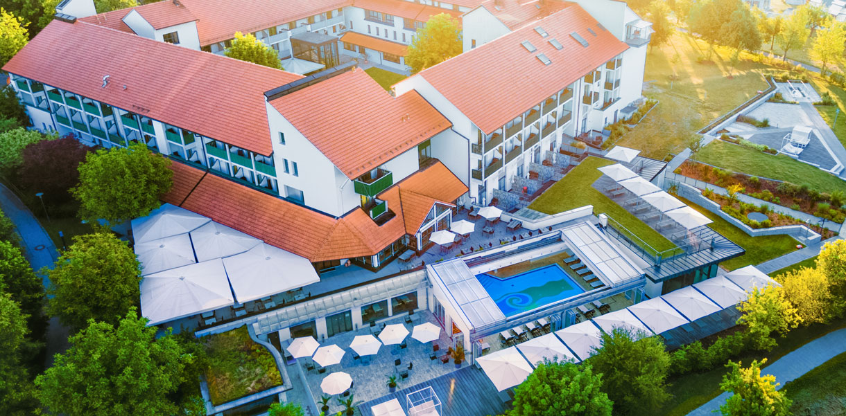 Eine Luftaufnahme des Hotel Sonnengut, eines eleganten Wellnesshotels mit roten Dachziegeln und moderner Architektur. Die Außenanlage umfasst Terrassen mit Sonnenschirmen, ein stilvolles Schwimmbecken und grüne Gärten. Die warme Abendsonne taucht die Szenerie in ein goldenes Licht.