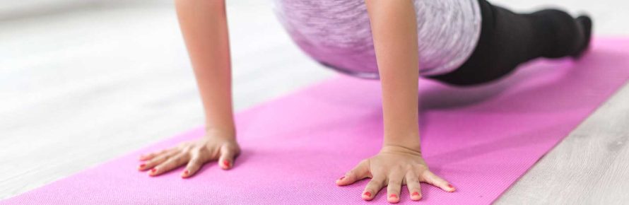 Eine Frau in Sportkleidung auf einer pinken Yogamatte in einem hellen Raum, in der Kindhaltung (Balasana) mit nach vorne ausgestreckten Armen und gesenktem Oberkörper.